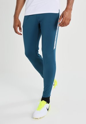Homme portant un pantalon de sport bleu avec des bandes blanches sur les côtés et des crampons blancs avec des lacets jaune néon et des chaussettes noires, debout devant un fond uni.