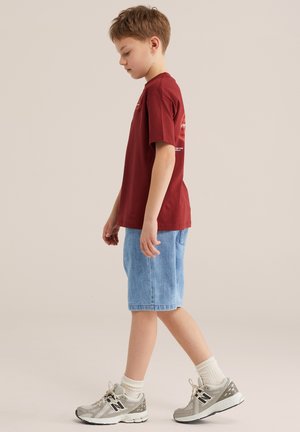 Poiss kõnnib, seljas punane lühikeste varrukatega särk, helesinised teksapüksid, valged sokid ja hallid tossud, taustaks neutraalne beež värv.