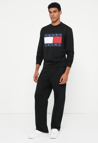 Mustal t-särk, millel on sinine, valge ja punane "TOMMY JEANS" logo, koos mustade pükste ja valgete jooksujalatsitega. Lihtne, kaasaegne disain.