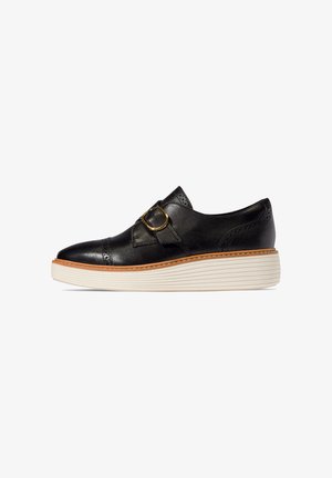 Scarpa slip-on in pelle nera con fibbia dorata, dettagli brogue e robusta suola piattaforma bianca rigata.