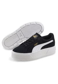 Zapatillas de ante negro con un diseño de franjas blancas y detalles dorados, que cuentan con una gruesa suela blanca y un patrón de textura en la suela.
