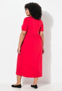 Robe rouge à manches courtes, longueur genou, avec un corsage ajusté et une taille froncée. Tissu lisse et coupe décontractée. Mocassins noirs portés.