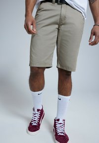 Shorts beiges en coton, arrivant aux genoux avec un ourlet droit. Portés avec des chaussettes blanches Nike et des baskets montantes bordeaux.