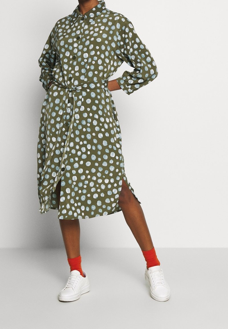 Robe verte à motifs avec des points blancs et bleu clair, boutonnée sur le devant, à manches longues, ceinture à la taille et fentes latérales ; associée à des baskets blanches.