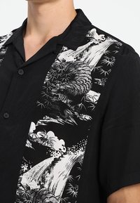 Zwarte korte mouwen shirt met een gedurfde witte graphic van een tijger, bamboe en golven aan de linkerkant. Knoopsluiting en een relaxte pasvorm.
