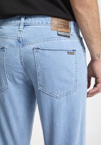 Jeans in denim blu chiaro con una patch in pelle sulla parte posteriore della vita, due tasche posteriori e un'etichetta logo discreta vicino alla tasca destra.