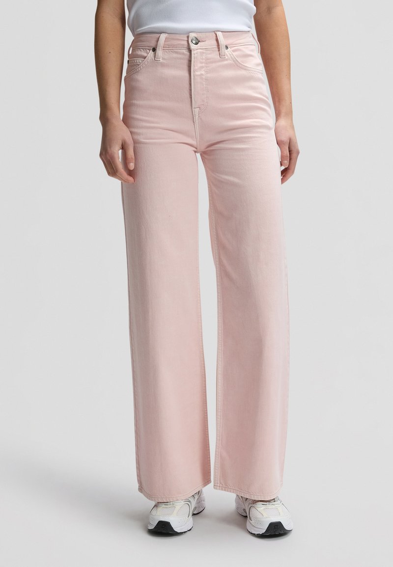 Γυναίκα που φοράει τζιν σε ανοιχτό ροζ χρώμα με ψηλή μέση και φαρδιά μπατζάκια, καθώς και λευκά αθλητικά παπούτσια μπροστά σε απλό γκρι φόντο.