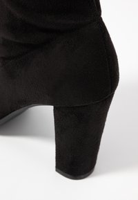 Bottine en suede noir avec un talon épais, présentant une texture lisse et un design sans couture. Accent sur la forme et le matériau du talon.