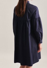 Námořnická modrá bavlněná šaty s volným střihem, nabíraným zadním dílem, dlouhými rukávy a knoflíkovými manžetami. Hladká textura a délka po kolena.