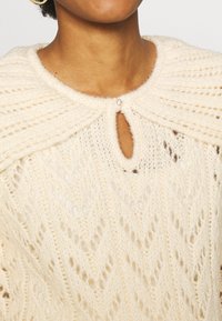 Krämfärgad stickad poncho med ribbat krage, med ett nyckelhål som säkras av en ädelstensknapp och ett texturerat spetsmönster.