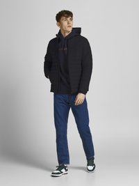 Giacca trapuntata nera sopra un felpa blu navy, jeans blu e sneakers verdi e bianche. Il modello è in piedi con un'espressione neutra su uno sfondo chiaro.