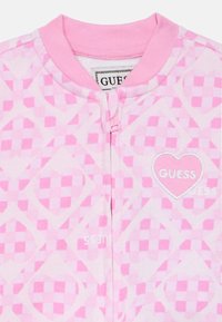 Indumento rosa con cerniera caratterizzato da un motivo a scacchi con cuori. Tessuto morbido e scollo a costine. Logo "GUESS" ricamato sul petto.