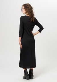 Vestido negro midi con mangas tres cuartos, que presenta una silueta ajustada y sutilezas en las costuras. Combinado con botines de tacón negros.