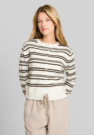 Junge Frau mit langen blonden Haaren, die eine weiße Strickjacke mit braunen horizontalen Streifen und beige Kordelhosen vor einem grauen Hintergrund trägt.