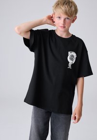 Fekete rövid ujjú pamut póló, melynek mellkasán grafikai nyomat található. Halvány szürke farmerrel párosítva. Egyszerű dizájn, kényelmes szabás.