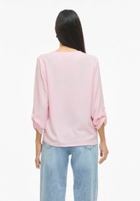 Femme aux longs cheveux foncés portant un chemisier rose clair à manches longues et un jean bleu clair, debout dos à l'appareil photo.