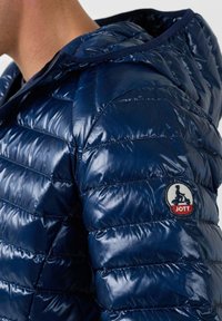 Marine pufferjack met glanzend, gewatteerd materiaal. Het ontwerp heeft een capuchon en een ronde logo-embleem met "JOTT" op de linkerschouder.