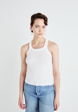 Top sans manches blanc à côtes avec un col rond, associé à un jean bleu clair. Texture lisse et design ajusté.