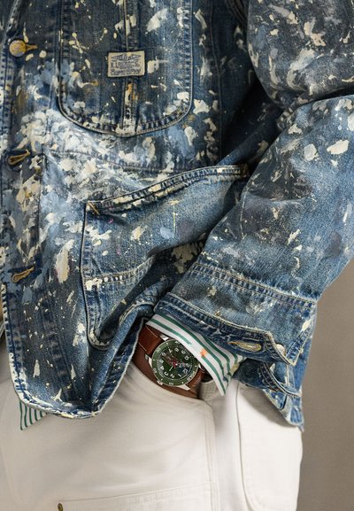 Άτομο που φοράει τζιν μπουφάν με κηλίδες μπογιάς, μανσέτα ριγέ πουκαμίσου, κρεμ παντελόνι και ρολόι χειρός με πράσινο καντράν και καφέ δερμάτινο λουράκι.