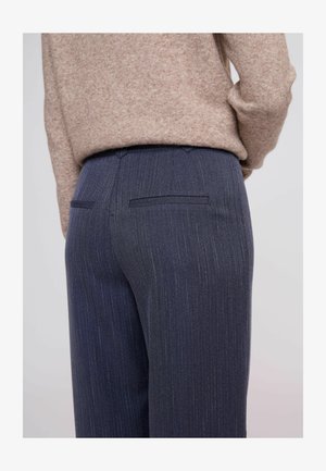 Lähellä kuva henkilöstä, joka käyttää tummansiniset raidalliset housut, joissa on takana kengurutaskut, ja beigen villapuseron, joka on tunkattu vyötärölle.