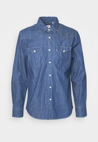 Long-sleeve denim shirt in dark blue with pointed yoke, two chest pockets, white buttons, and contrast stitching. Smooth texture.