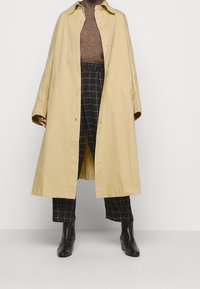 Beige bomull trenchcoat med knäppning fram, vida ärmar och sidofickor, kombinerad med svart och vit rutiga byxor och svarta ankelstövlar.