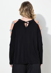 Schwarzes Oberteil mit kaltem Schulterdesign, ausgestattet mit silbernen Akzentträgern und einer Bindeschließe auf der Rückseite. Hergestellt aus weichem, fließendem Stoff.