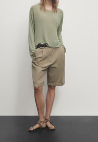 Lichtgroene langarmtop, khaki geplooide shorts, zwarte riem en bruine sandalen met banden, tegen een neutrale achtergrond.