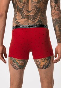 Boxers rouges avec des motifs en losanges noirs, taille élastique arborant "JP1880," vus de l'arrière, mettant en valeur des jambes tatouées.