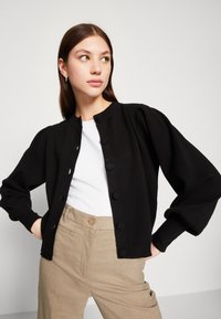 Zwart gebreid cardigan met poffige mouwen en een ronde halslijn, voorzien van gekleurde knopen en gedragen over een witte geribbelde top.