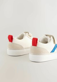 Białe i beżowe sneakersy z czerwoną pętlą na pięcie, niebieskim paskiem po bokach i teksturowanymi zamszowymi akcentami. Okrągły kształt i gumowe podeszwy.