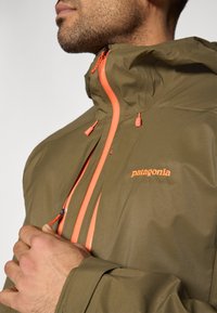 Αδιάβροχο μπουφάν σε ελιά πράσινο με ρυθμιζόμενο κουκούλα, πορτοκαλί φερμ ουάρ και λεπτομέρειες, με τσέπη με φερμουάρ μπροστά και λογότυπο στο στήθος.