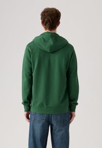 Felpa verde realizzata in un tessuto morbido, con cappuccio regolabile, polsini a coste e orlo dritto, abbinata a jeans di denim blu.
