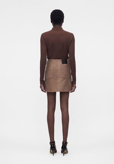 Γυναίκα που στέκεται με την πλάτη, φοράει ένα καφέ ριμπ μπλουζάκι με μακριά μανίκια, μίνι δερμάτινη φούστα σε ταμπ απόχρωση με τσέπες πίσω και μαύρα παπούτσια με ψηλά τακούνια.