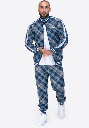 Blauer Karosportanzug aus leichtem Material mit weißen Akzenten, bestehend aus einer zip-up Jacke und elastischen Bündchen, kombiniert mit weißen Sneakern.