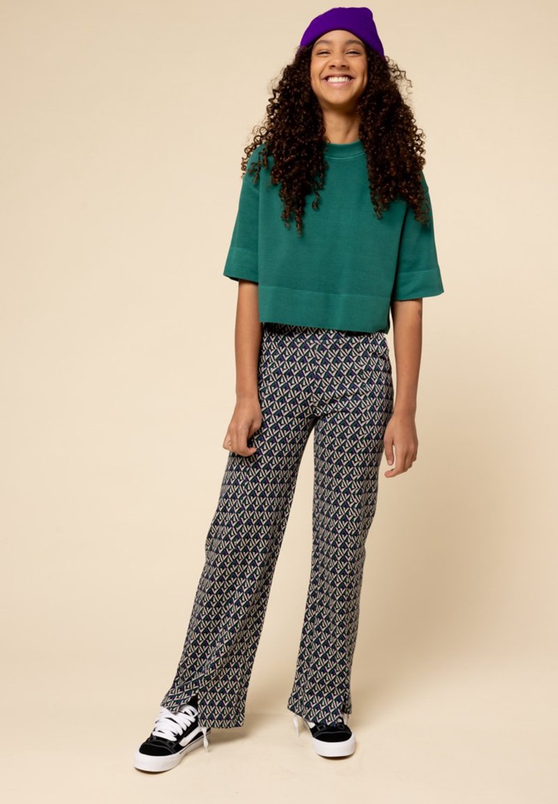 Groene cropped sweatshirt, wijde broek in marineblauw met een geometrisch patroon en zwarte sneakers met witte zolen. Paarse beanie op het hoofd.