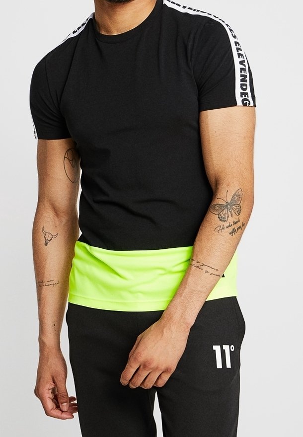 Homme portant une chemise noire à manches courtes avec un texte blanc sur les épaules et un accent jaune néon, associé à un pantalon noir avec un logo blanc 11°.