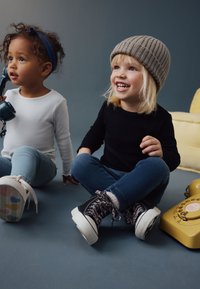 To børn sidder på en blå gulv. Den ene har en hvid skjorte og lyseblå jeans på, den anden har en sort skjorte og blå jeans, med en ribbet beige hue og leopardmønstrede sko.