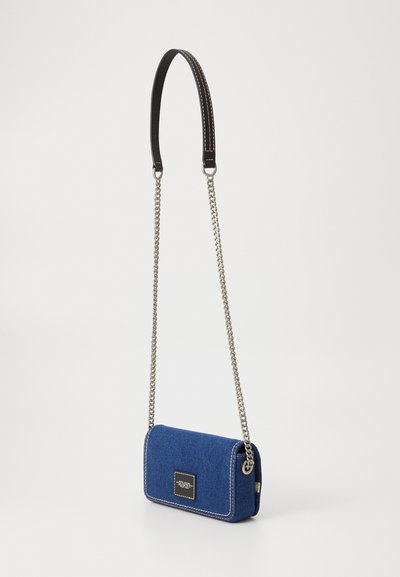 Geantă din denim cu bretea crossbody, de culoare albastră, cu cusături albe contrastante, având un șnur de lanț argintiu, accente din piele neagră și un patch cu logo pătrat.