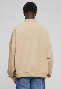 Osoba s krátkými kudrnatými vlasy má na sobě oversized béžovou fleece mikinu a modré džíny, stojí zády k jednofonnímu světle šedému pozadí.