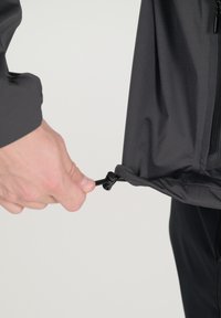 Zwarte buitenkleding met een gladde textuur heeft een koord aan de onderrand, dat door een hand wordt aangepast. Het ontwerp is gestroomlijnd en functioneel.