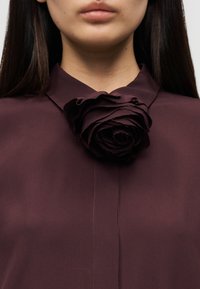 Μπλούζα μπορντό με λουλούδι από υφασμάτινη υφή και πολλαπλά στρώματα στο γιακά. Λείο ύφασμα, κλείσιμο με κουμπιά, χωρίς ορατά σχέδια ή μεταλλικά μέρη.