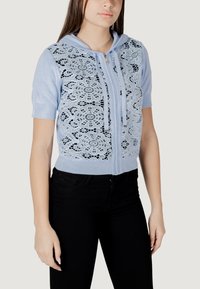 Sudadera corta azul claro con detalles de encaje en la parte frontal, mangas cortas y cremallera delantera completa. Se llevan jeans negros abajo.