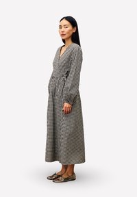 Femme se tenant de côté, portant une robe à manches longues à carreaux noir et blanc et des ballerines en bronze métallique sur un fond blanc.