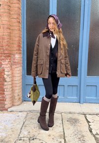Brun isolerad jacka med fickor, svart kofta, leggings, bruna knähöga stövlar och en grön handväska med en plyschaccessoar. Lila scarf.