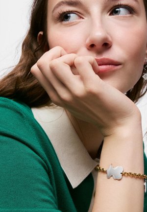 Young woman with hand near face wearing green knit top with white collar and gold bracelet featuring a silver teddy bear charm.