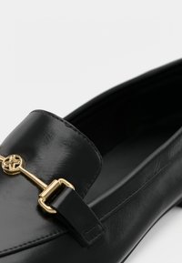 Mocassin en cuir noir avec une finition brillante, doté d'un détail en métal doré et d'une doublure intérieure lisse.