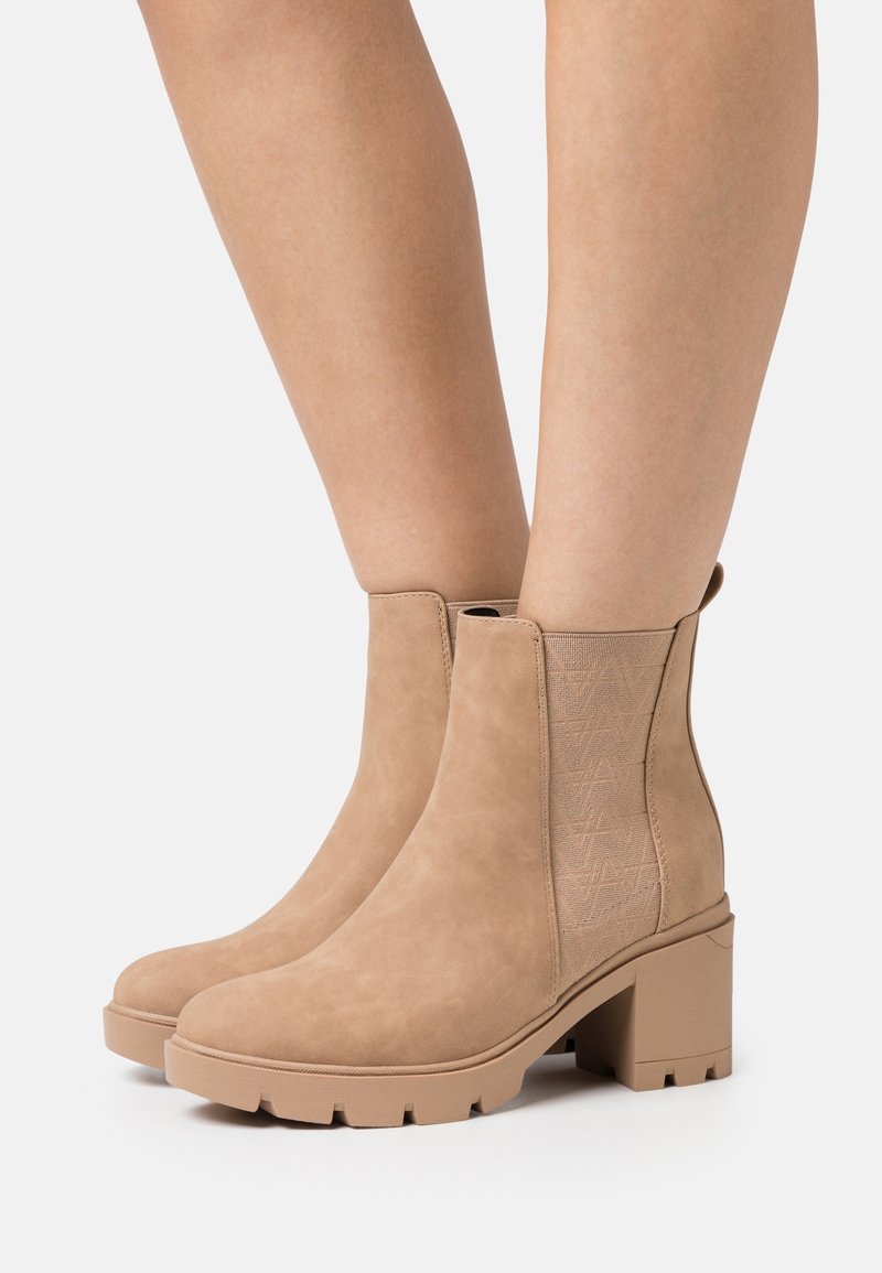 Bottes basses en daim beige avec talons épais et panneaux latéraux élastiques portées sur des jambes nues sur un fond clair.