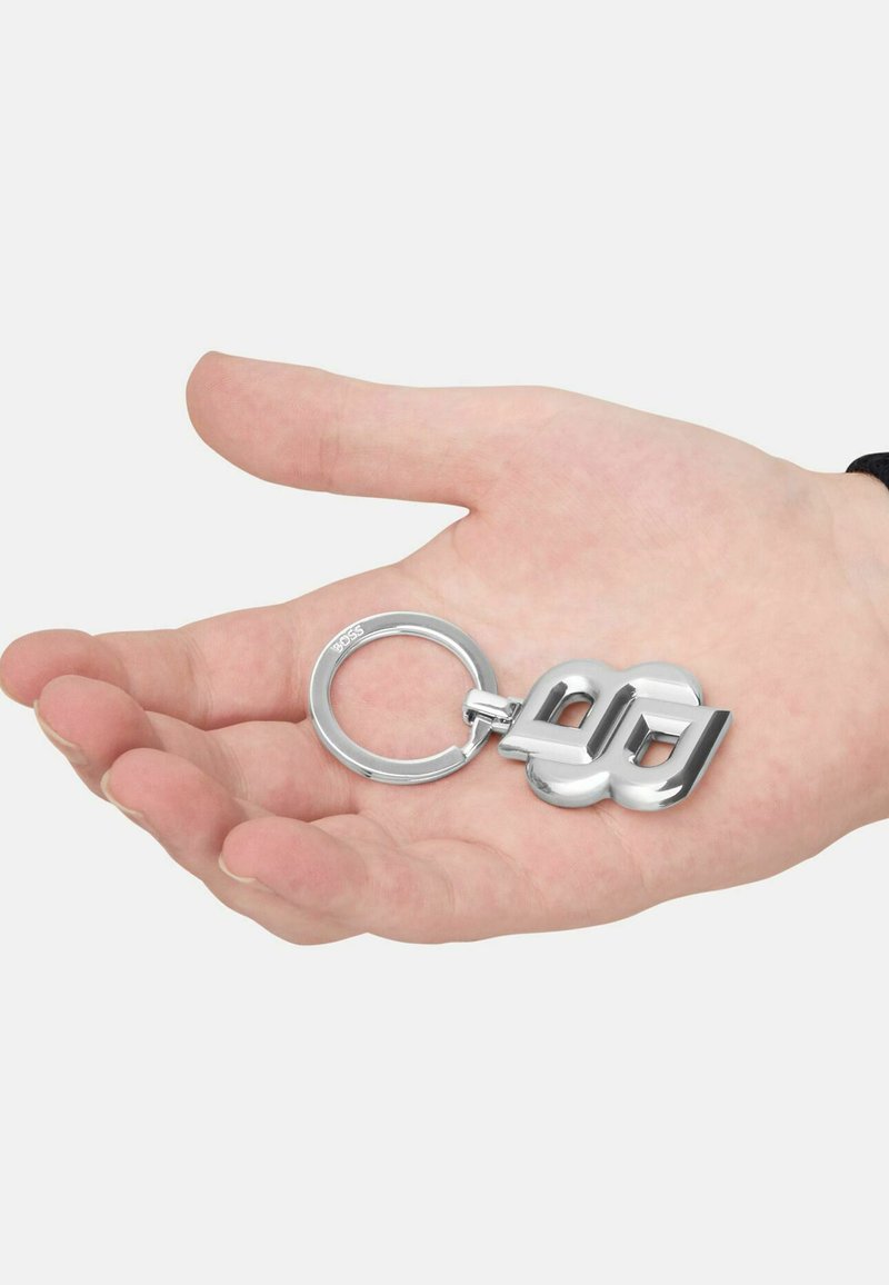 Silver metal keychain with a modern design featuring linked letters, held in an open palm against a plain background.