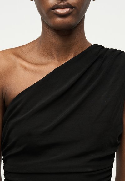 Κοντινό πλάνο ενός ατόμου που φοράει μαύρο μπλουζάκι με έναν ώμο, εμφανίζοντας τον λαιμό, τα χείλη και τα σκουλαρίκια σε απλό φόντο.